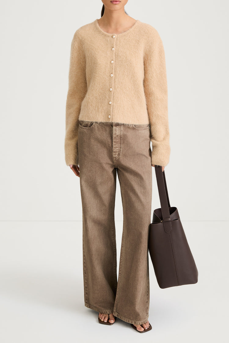 Model wearing a beige cardigan and brown pants, holding a brown bag on a white background