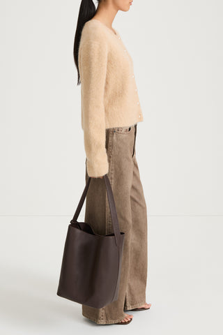 Model wearing a beige sweater and brown pants holding a dark brown bag on a white background