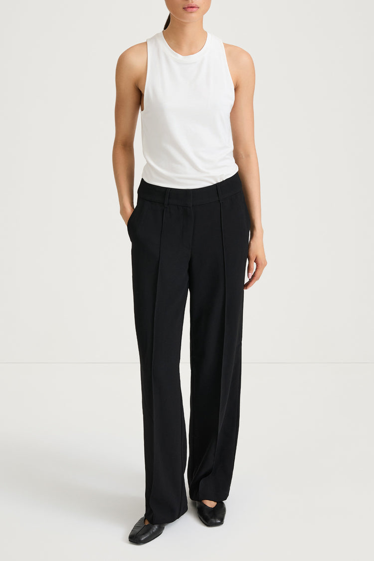 A model wearing black wide-leg pleated trousers with a white sleeveless top, standing against a plain white studio background.