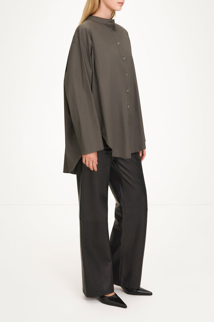 A model wearing an oversized, high-neck button-front top in dark olive gray with wide-leg black trousers and black pointed shoes, photographed against a clean white studio background.