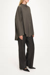 A model wearing an oversized, high-neck button-front top in dark olive gray with wide-leg black trousers and black pointed shoes, photographed against a clean white studio background.