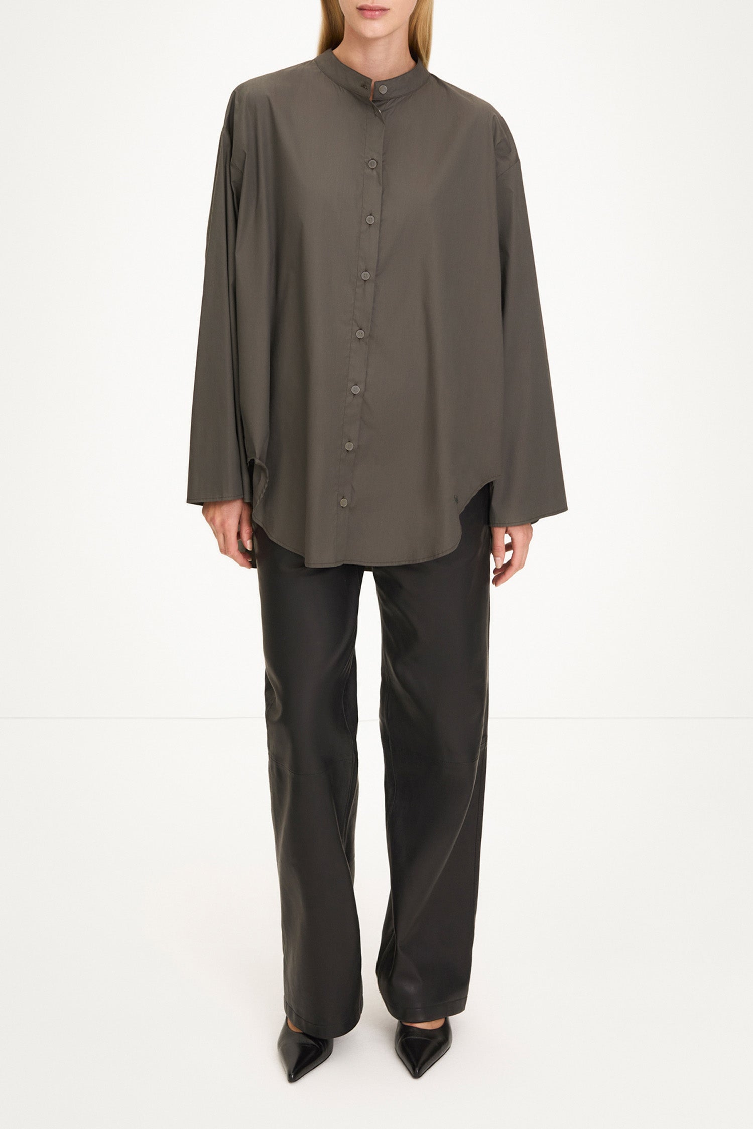 A model wearing a dark olive brown long-sleeve button-up shirt with a high mock neck, paired with black wide-leg trousers and black pointed shoes, shot against a plain white studio background.