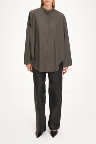 A model wearing a dark olive brown long-sleeve button-up shirt with a high mock neck, paired with black wide-leg trousers and black pointed shoes, shot against a plain white studio background.
