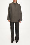 A model wearing a dark olive brown long-sleeve button-up shirt with a high mock neck, paired with black wide-leg trousers and black pointed shoes, shot against a plain white studio background.