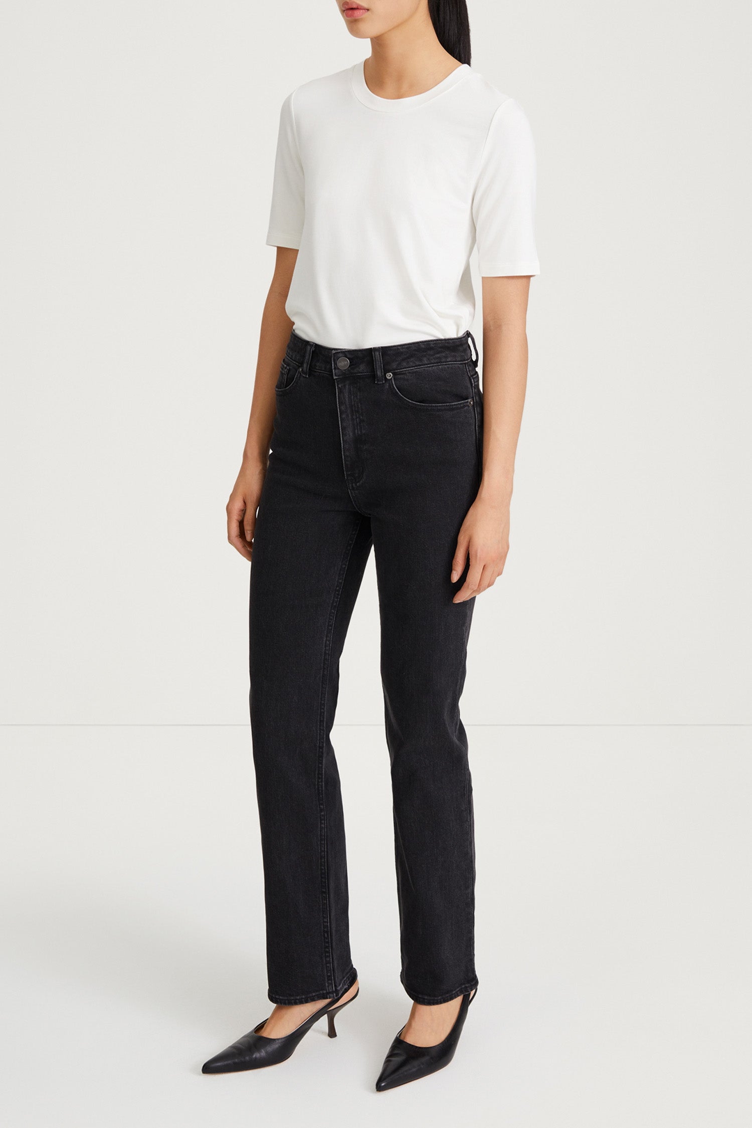 A model wearing a white short-sleeve crewneck t-shirt, black high-waisted straight-leg jeans, and black pointed-toe mule heels, standing against a plain white studio background.