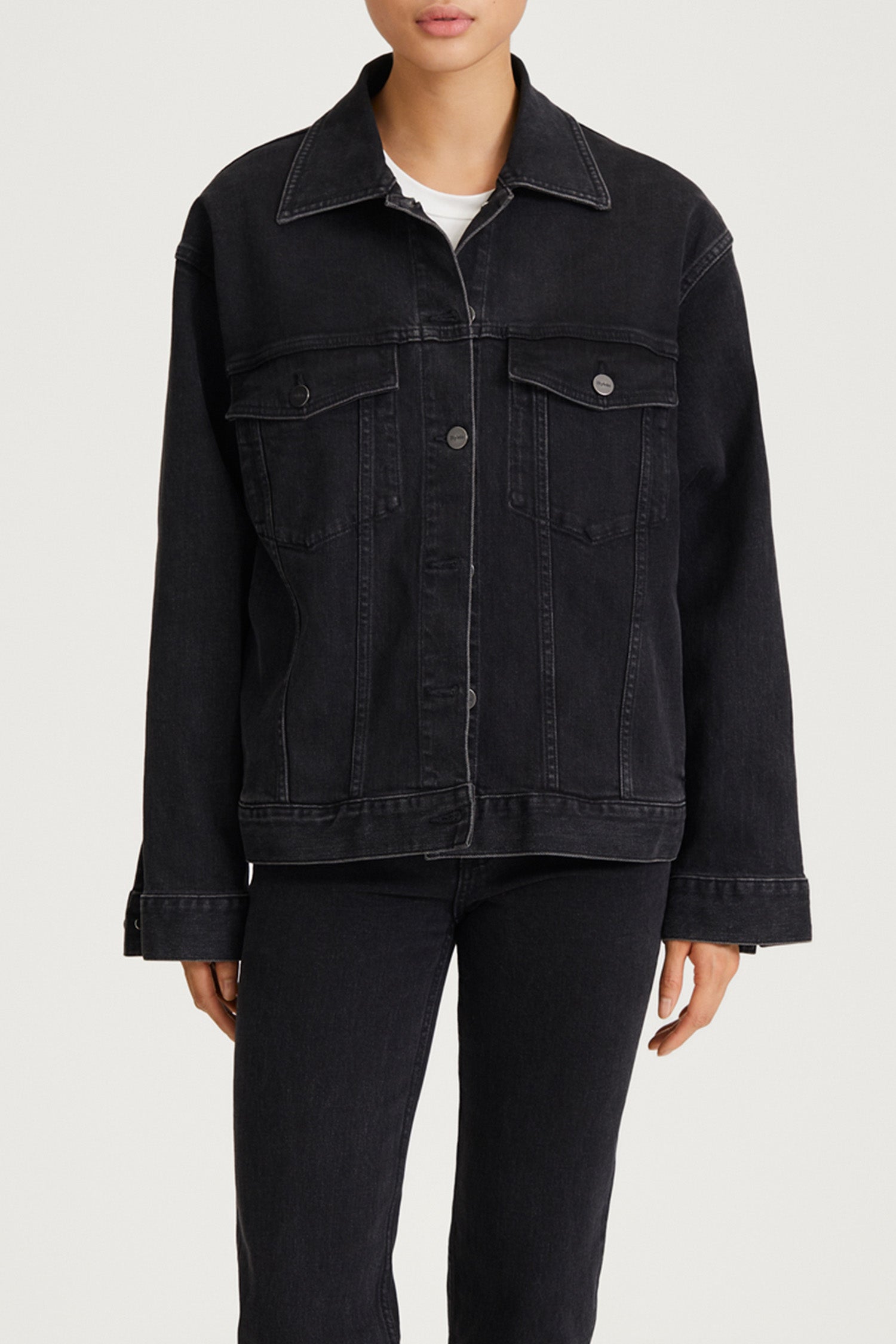 A model wearing a dark washed denim jacket with front flap pockets and button snaps, paired with dark jeans and a white undershirt, shown against a light gray studio background.