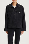 A model wearing a dark washed denim jacket with front flap pockets and button snaps, paired with dark jeans and a white undershirt, shown against a light gray studio background.