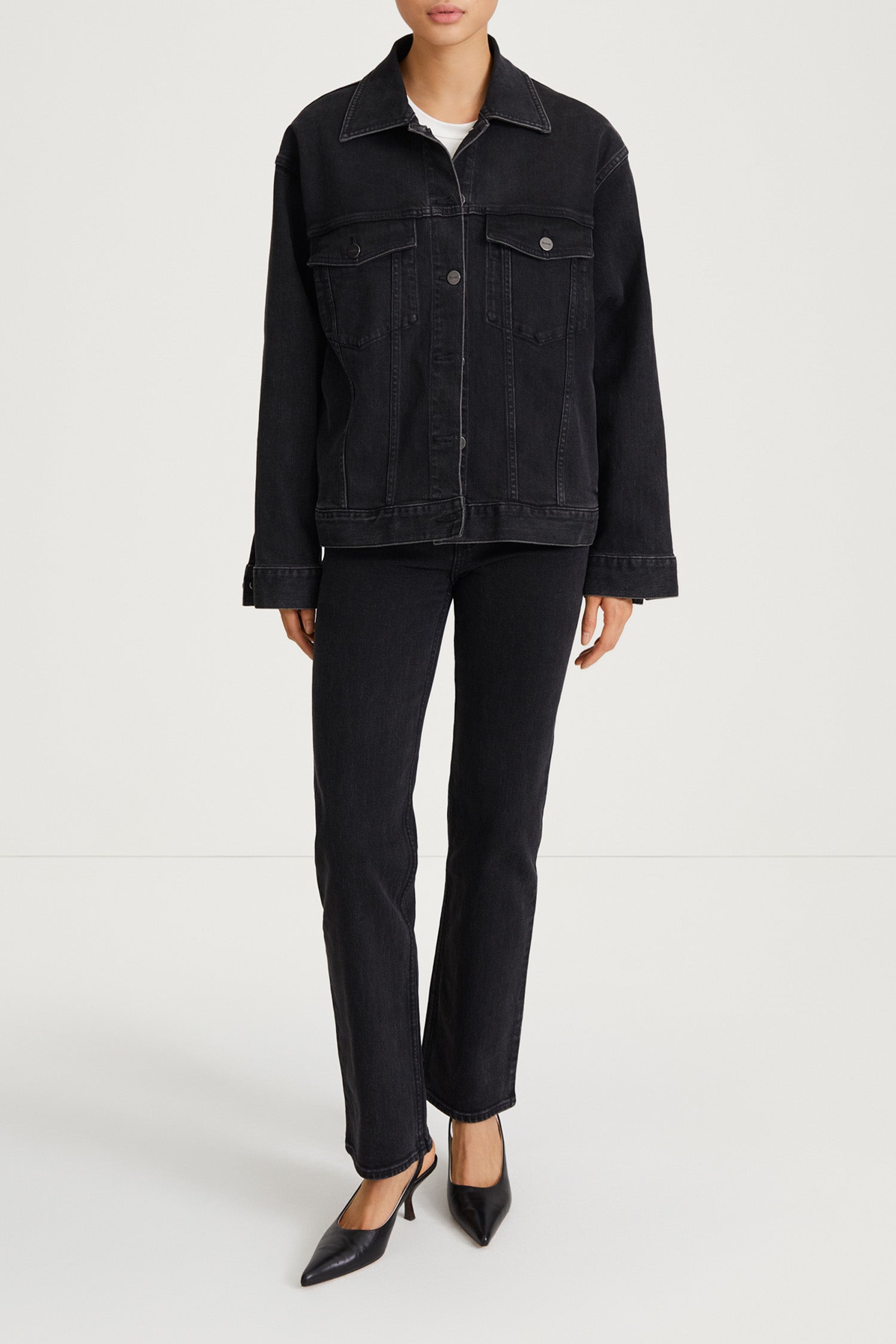 A model wearing a black denim jacket and matching black denim trousers, styled with black pointed-toe heels, against a plain light gray studio background.