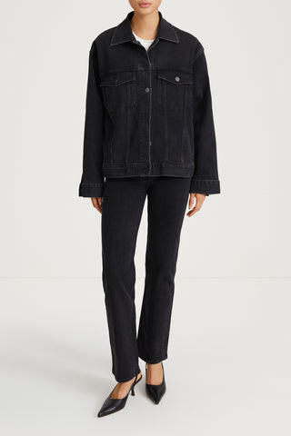A model wearing a black denim jacket and matching black denim trousers, styled with black pointed-toe heels, against a plain light gray studio background.