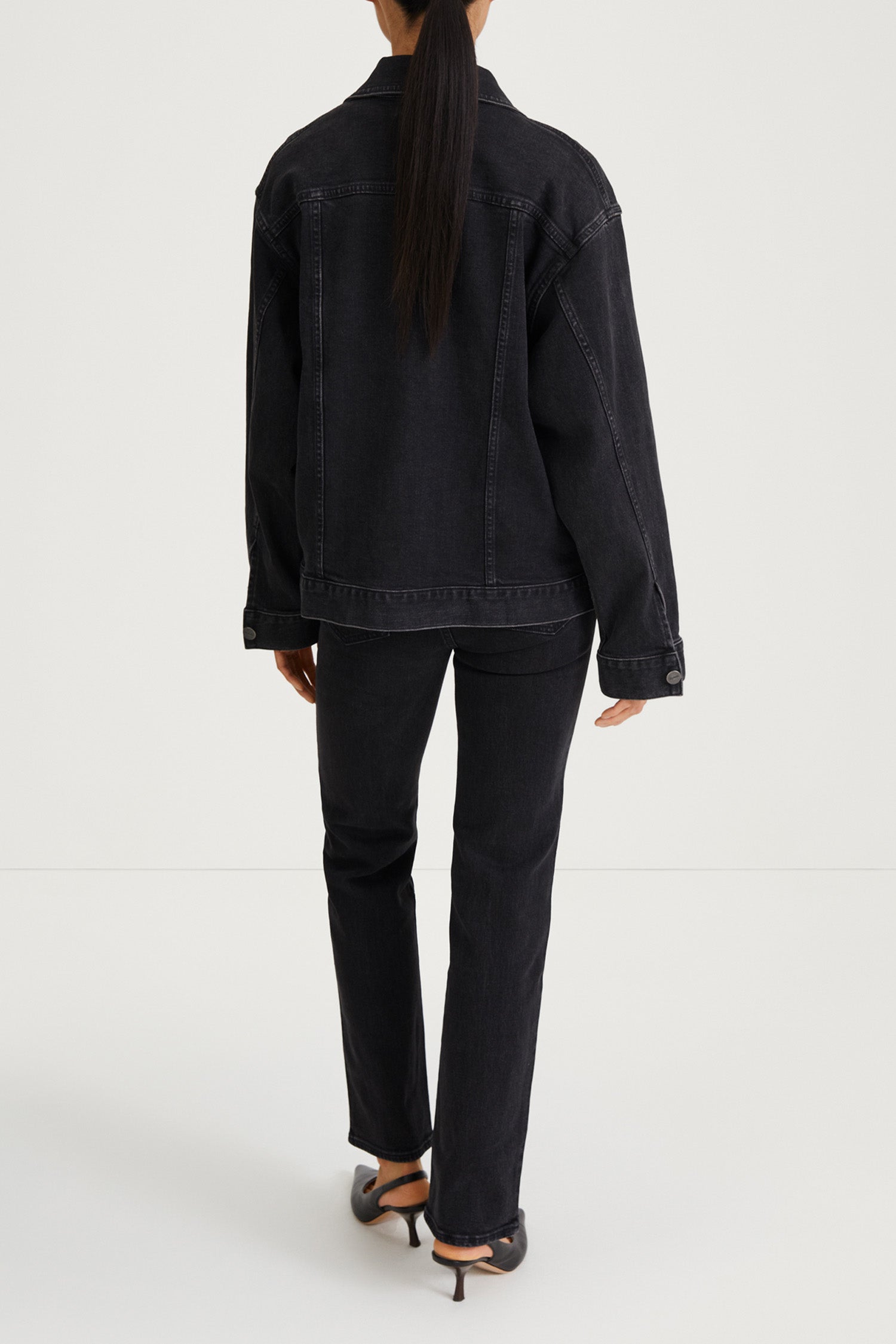 A model shown from the back wearing a black denim jacket and matching black jeans, paired with black high-heeled sandals, against a light gray studio background.