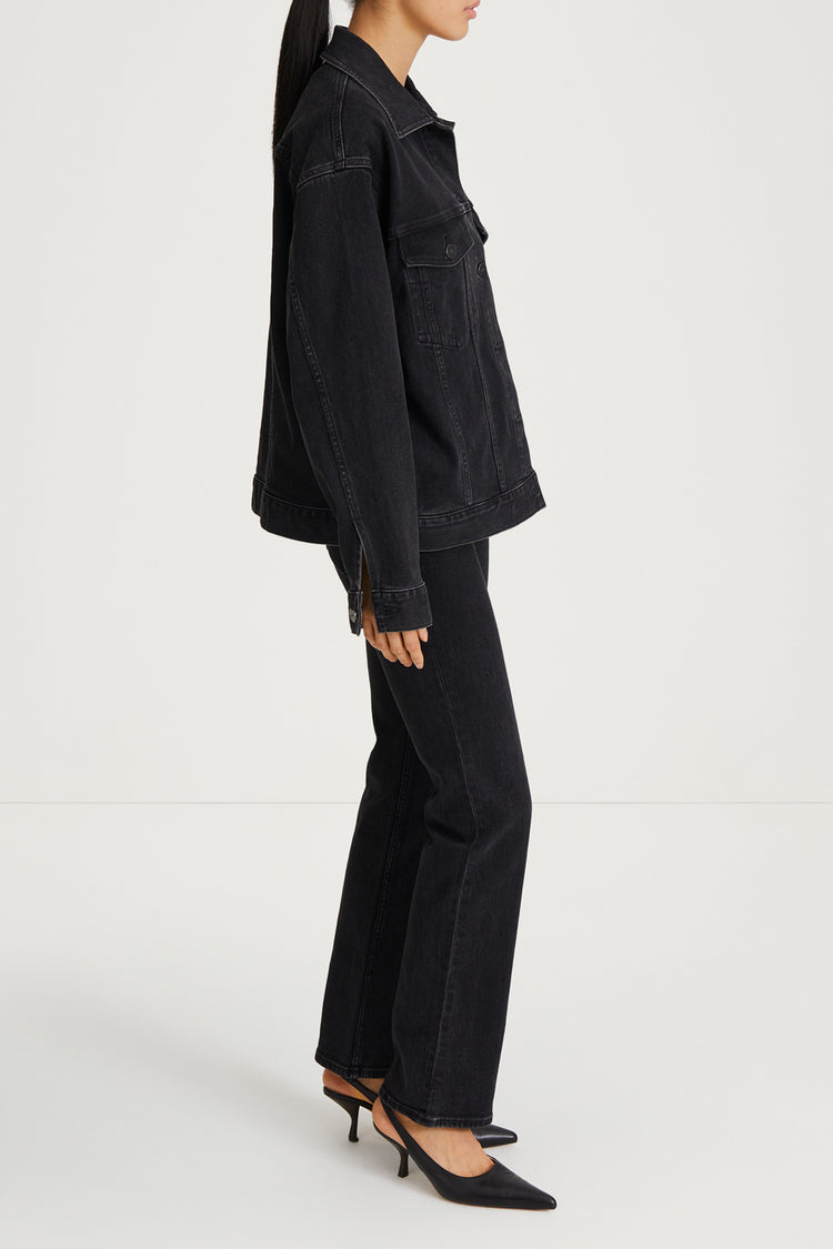 A model shown from the side wearing a black denim jacket and matching black wide-leg jeans, paired with black pointed-toe mule heels, against a plain light gray studio background.
