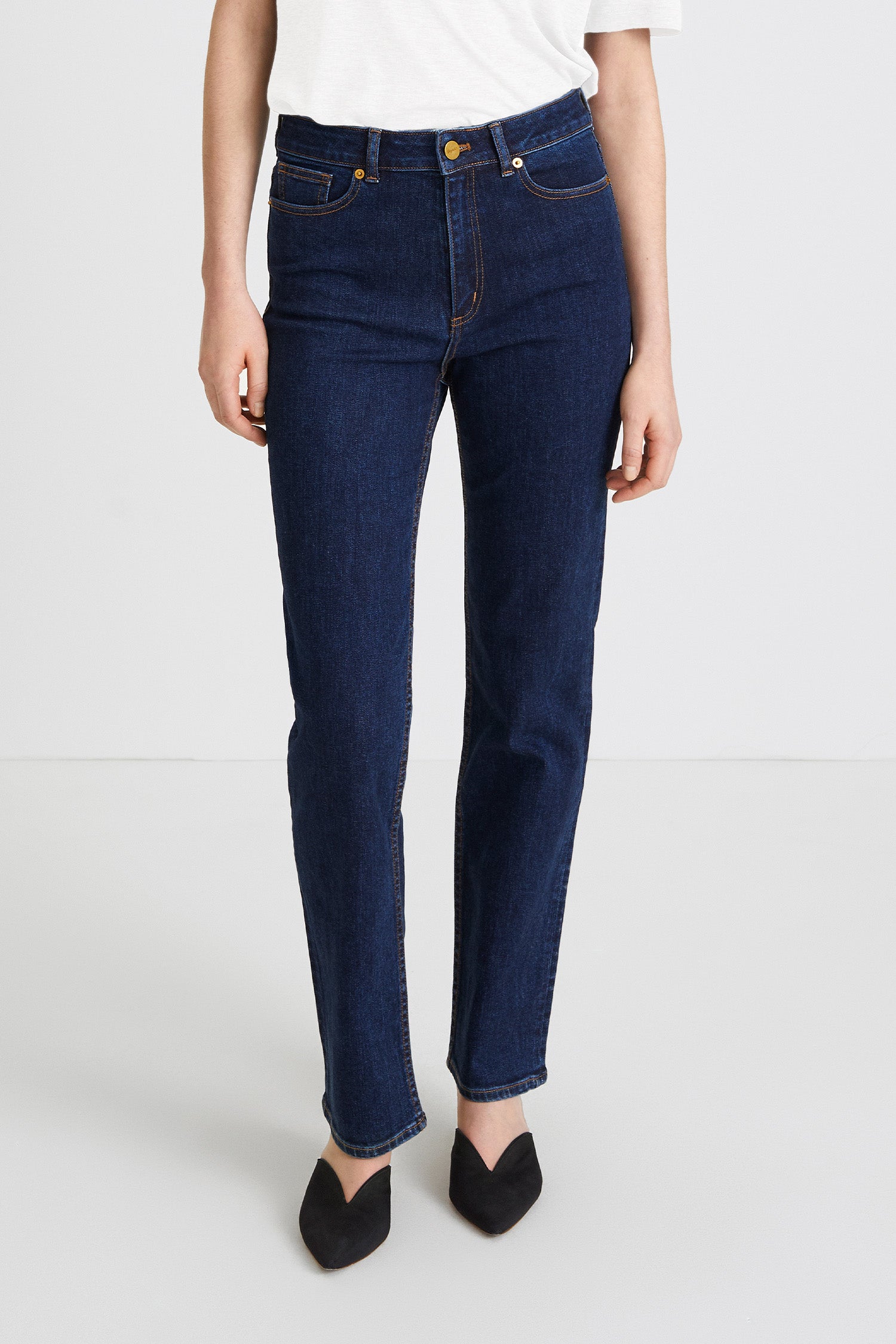 A model wearing dark blue straight-leg denim jeans, styled with a white t-shirt and black pointed-toe flats, photographed against a clean white studio background.