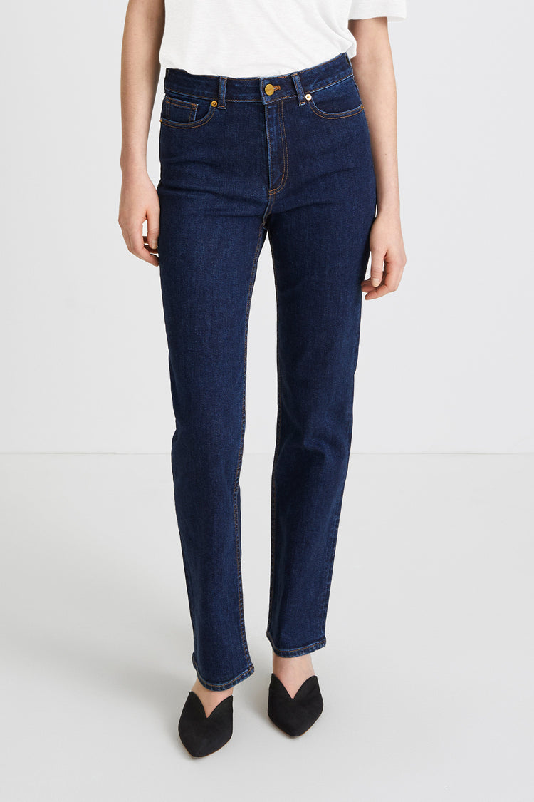 A model wearing dark blue straight-leg denim jeans, styled with a white t-shirt and black pointed-toe flats, photographed against a clean white studio background.