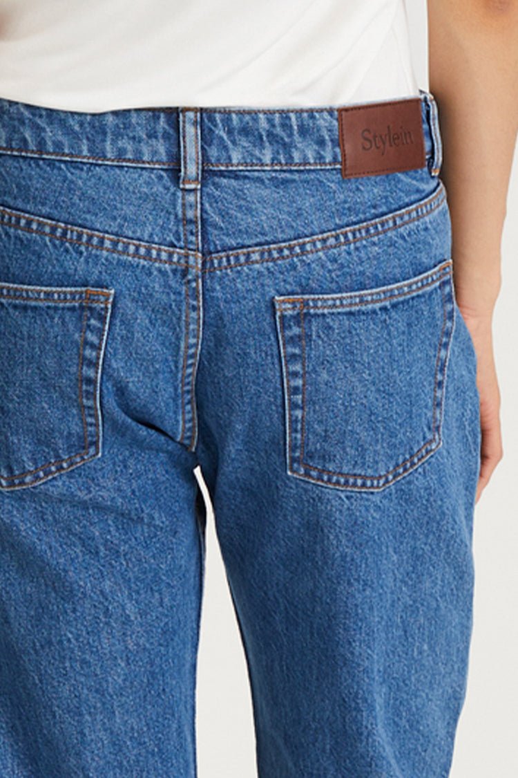 Close-up back view of a person wearing blue denim jeans with a white top, showing the waistband, belt loop, back pockets, and a brown leather patch with the brand name 