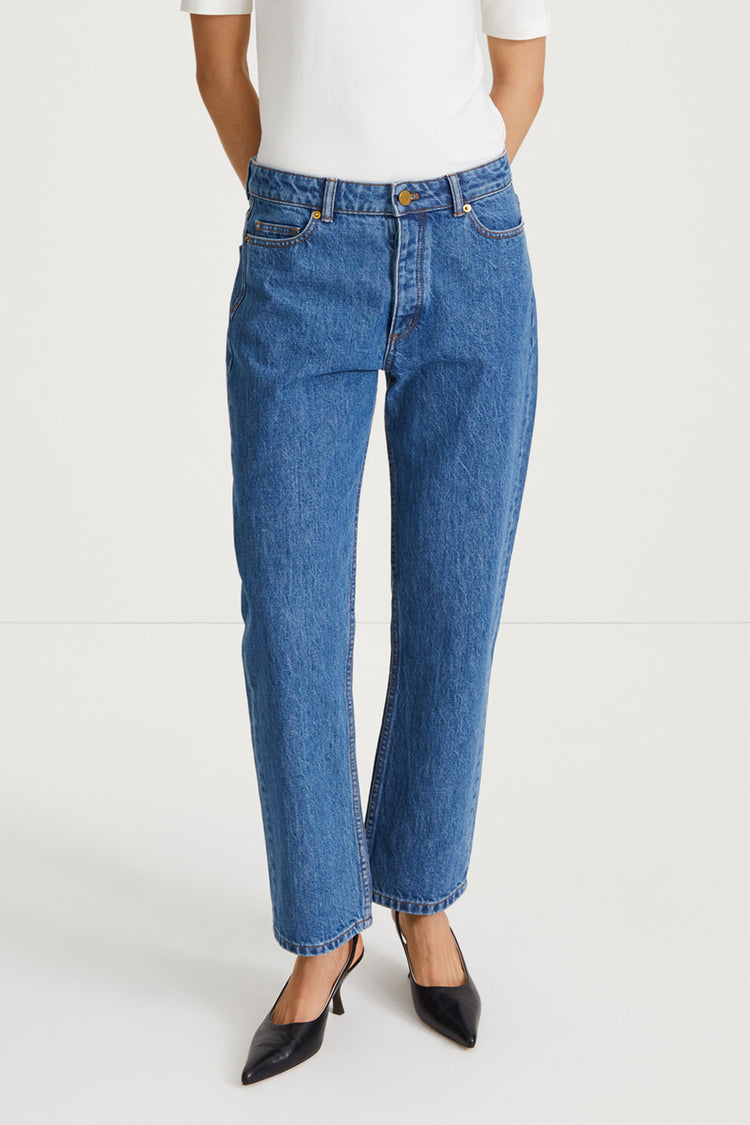 A model wearing a pair of medium-wash blue denim jeans with a straight-leg fit, paired with a white top and black pointed-toe heels, shown against a plain white studio background.