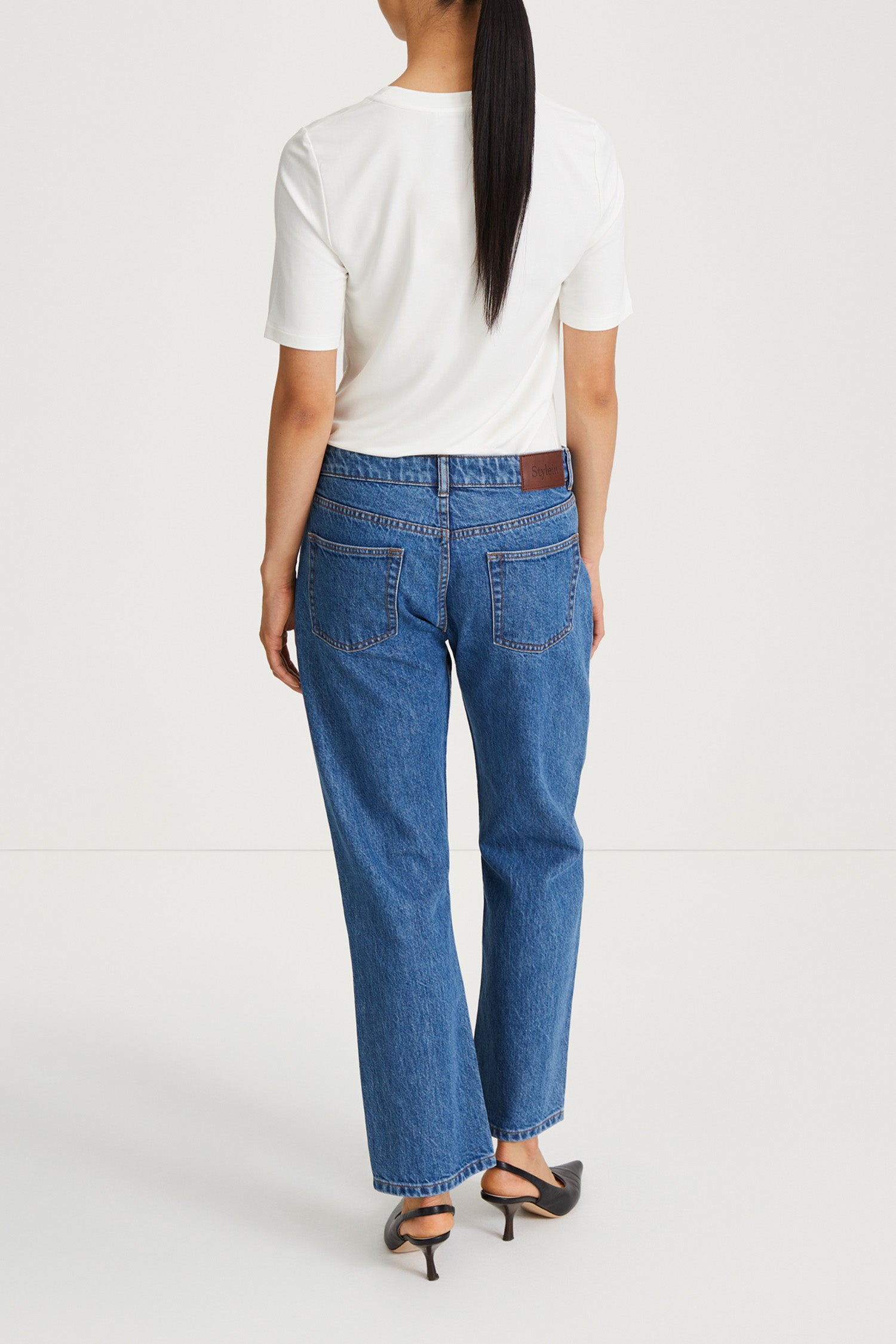 A woman viewed from behind wearing blue straight-leg jeans, a white short-sleeve t-shirt, and black heeled sandals against a plain light gray studio background.