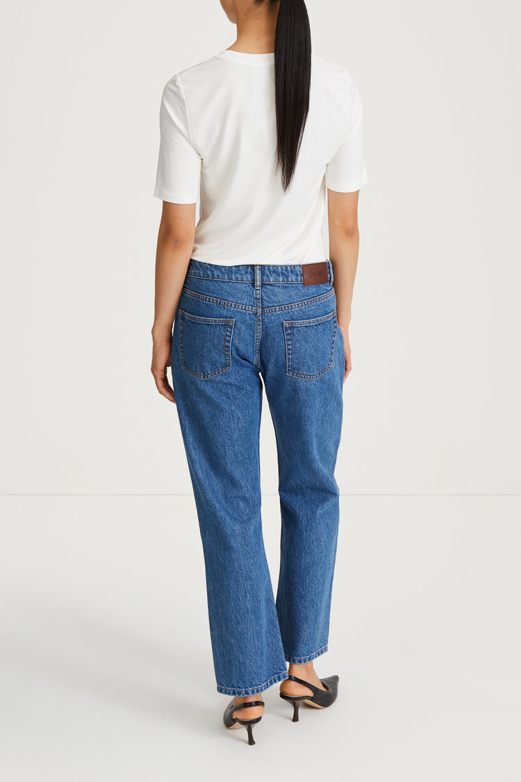 A woman viewed from behind wearing blue straight-leg jeans, a white short-sleeve t-shirt, and black heeled sandals against a plain light gray studio background.