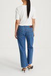 A woman viewed from behind wearing blue straight-leg jeans, a white short-sleeve t-shirt, and black heeled sandals against a plain light gray studio background.