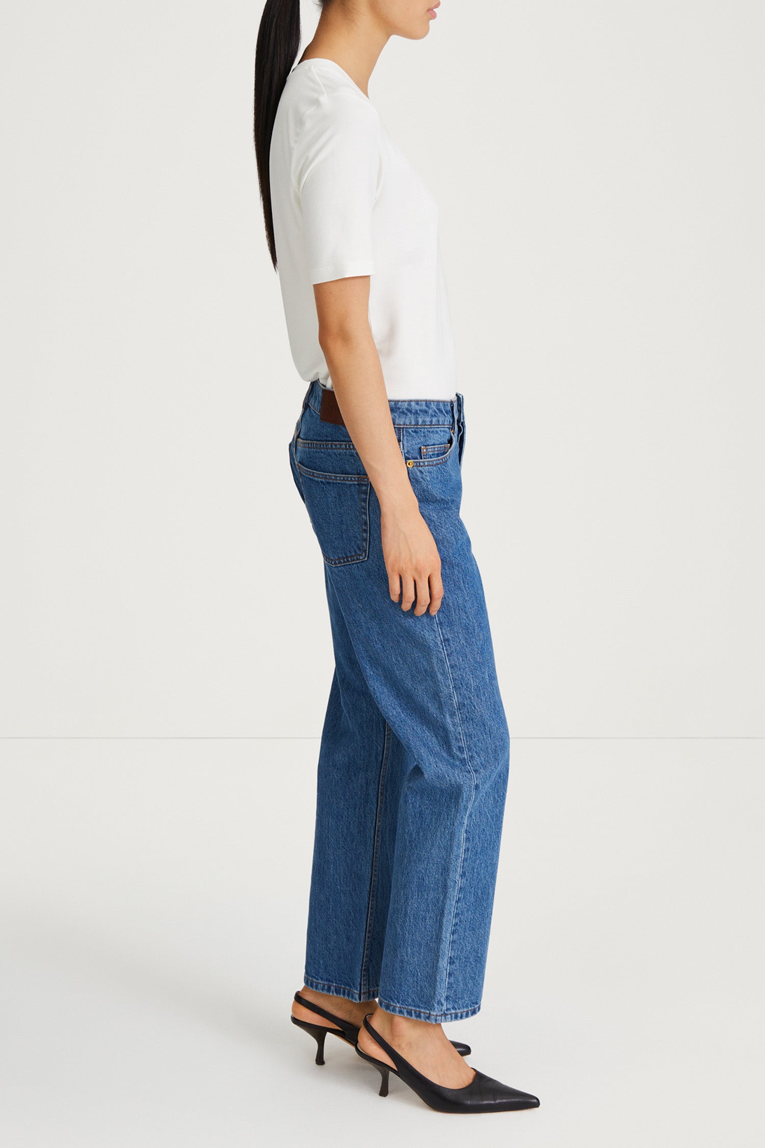 A side-profile view of a model wearing blue wide-leg cropped jeans, a white short-sleeve top, and black slingback heels against a clean white studio background.